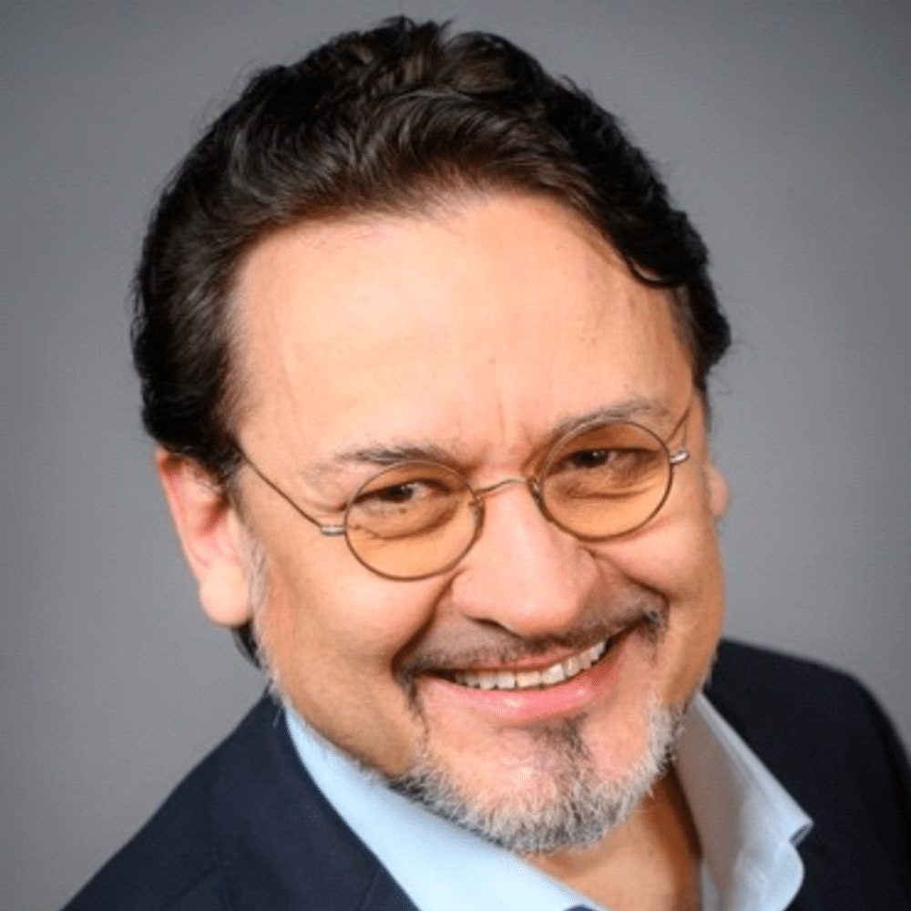 Smiling man with dark hair, round glasses, and a goatee wearing a dark blazer and light blue shirt.