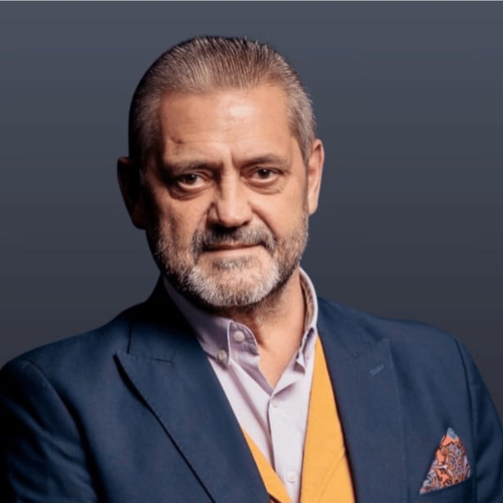 Man with grey hair and beard wearing a blue blazer, yellow vest, and patterned pocket square.